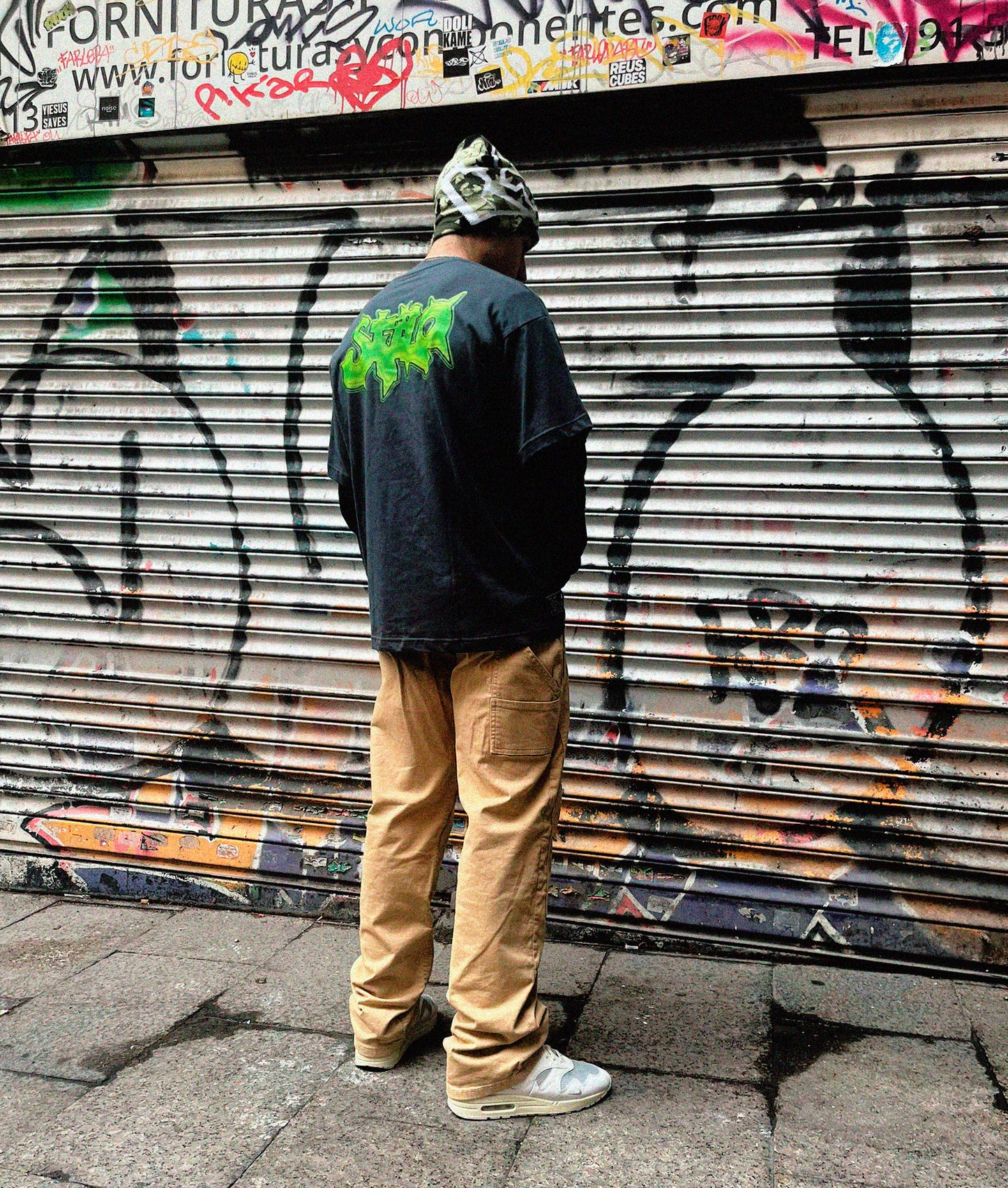 urban guy posing in front of a graffiti wall, wearing a grey and green t-shirt, hip-hop outfit.